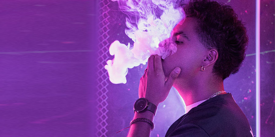 man with curly short black hair vaping and exhaling smoke in purple background