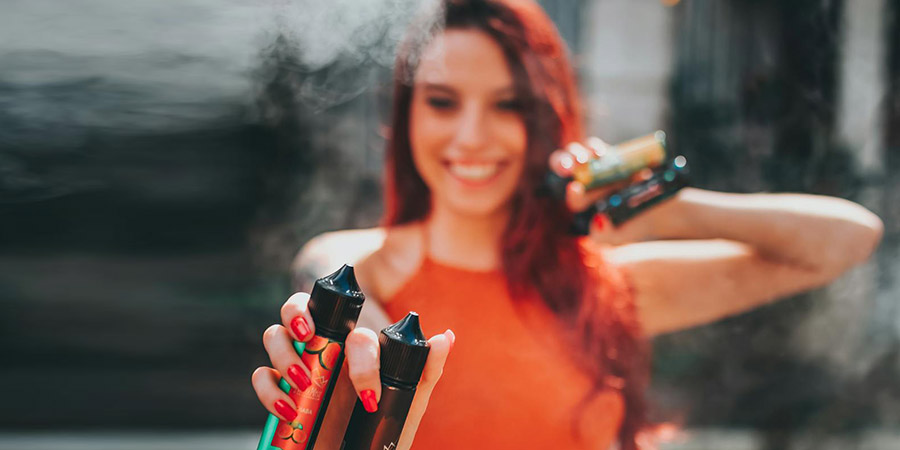 woman with red long wavy hair wearing orange top holding three vape juices in both hands